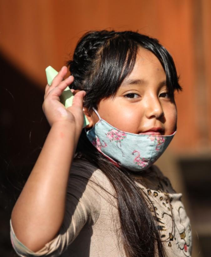 A child with long hair wears a decorative face mask under their chin, raises a hand holding a green object, and stands in a sunlit outdoor area with a blurred background.