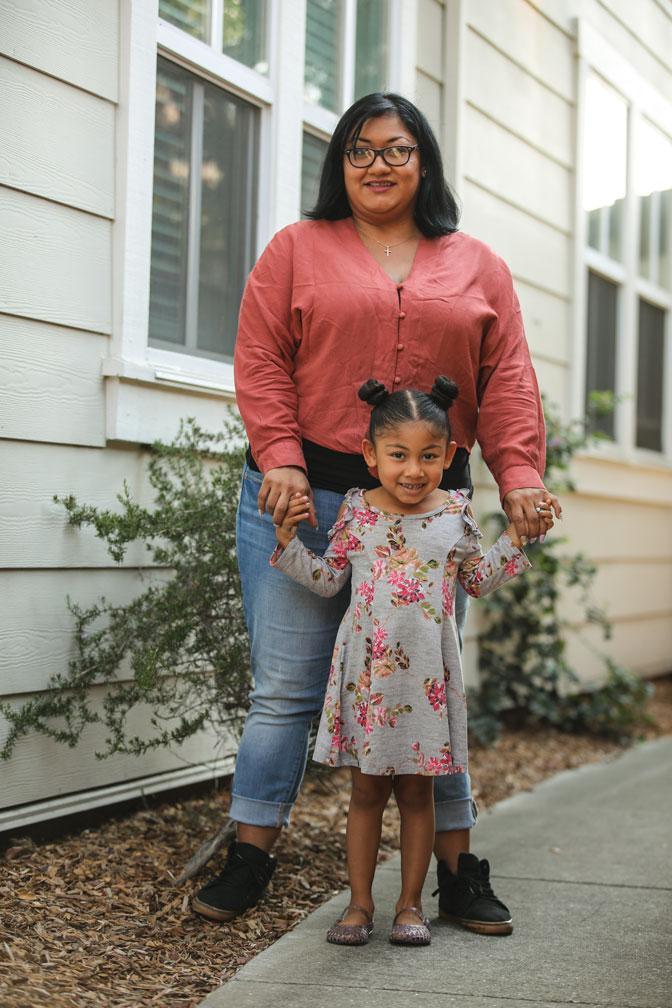 A woman wearing glasses and a red blouse stands holding hands with a young girl in a floral dress on a sidewalk beside a house with white siding and windows.