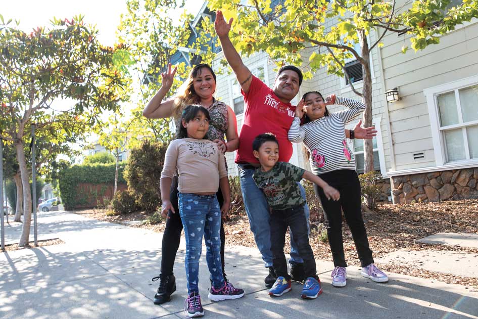 A family of five poses energetically on a sunlit residential sidewalk, surrounded by trees and apartment buildings. The man wears a red T-shirt with white text partially visible: "THERE IS..."