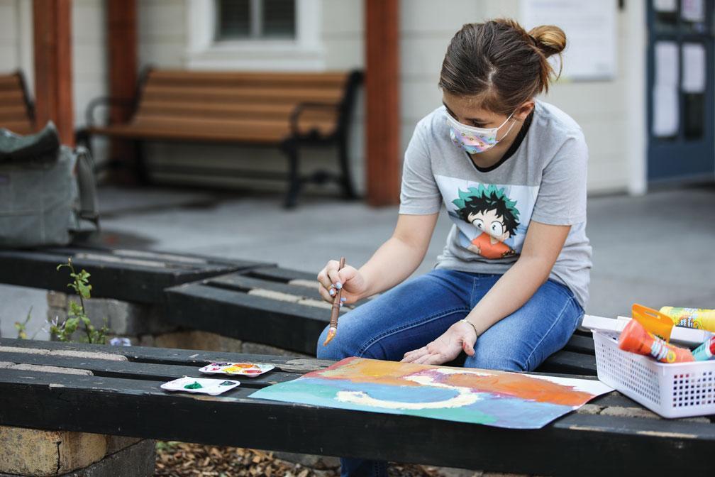 A child wearing a mask paints with a brush on paper while sitting on a bench in an outdoor setting, surrounded by art supplies.