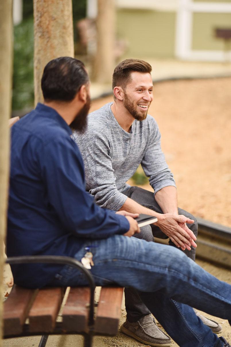 Two men sit on a bench, engaged in conversation, at an outdoor park with trees and a sandy ground. One holds a notebook.
