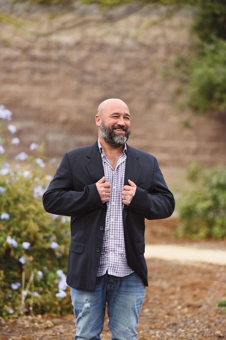 A smiling man adjusts his jacket while standing on a garden pathway, surrounded by blooming flowers and greenery, with a stone wall in the background.