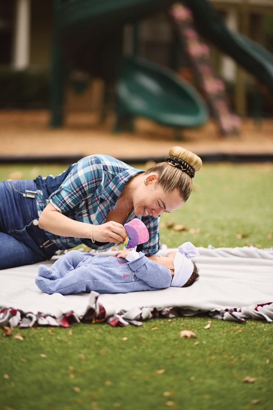 Woman playfully interacts with a baby holding a toy, on a blanket in a park setting. A playground slide is visible in the background.