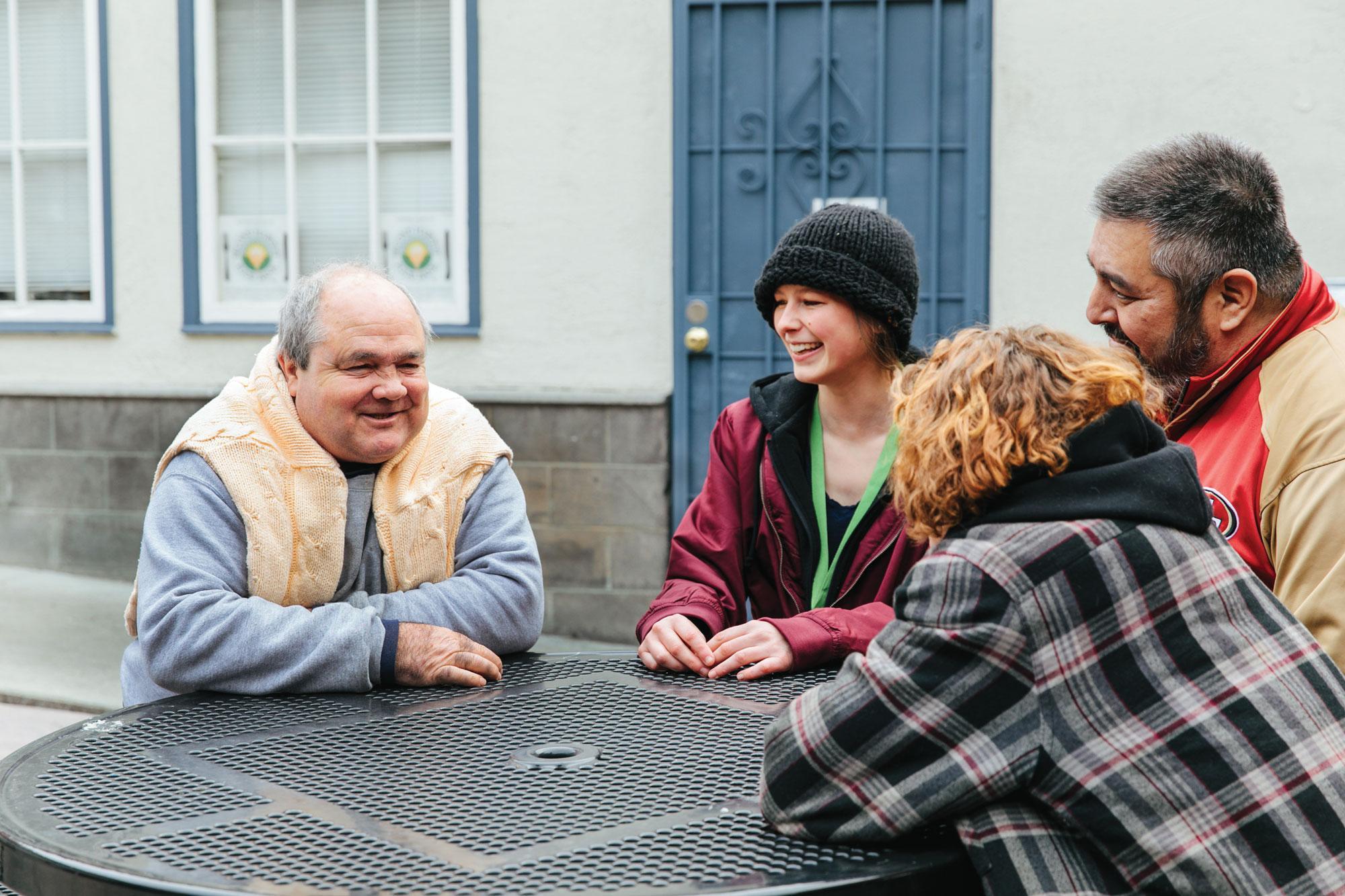 Four people seated around an outdoor metal table, engaged in a lively conversation. They are wearing casual, warm clothing, with a building featuring windows and a door in the background.