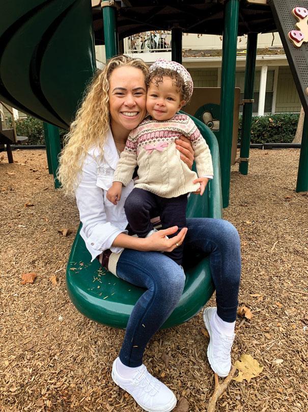A woman, smiling, sits on a green slide holding a child in a playground. The ground is covered with wood chips, and play structures are visible in the background.