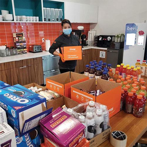 A person wearing a mask holds an orange Chegg box labeled "Fully booked with great stuff." in a kitchen with tables filled with snacks, drinks, and other Chegg boxes.