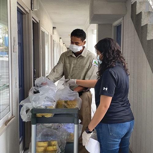 Two people, wearing face masks and gloves, handle packaged food on a trolley in a building corridor, preparing for distribution or delivery.
