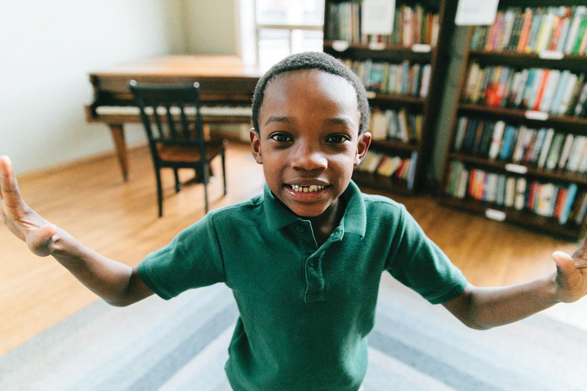 Child smiling with arms outstretched, standing in a room with wooden floors. Bookshelves filled with colorful books line the walls, and a piano with a chair is in the background.