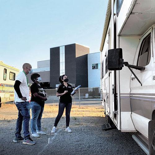 Three people stand beside a parked RV, one holding a clipboard, likely discussing or inspecting it. They're in a gravel lot outside a modern, multi-story building.