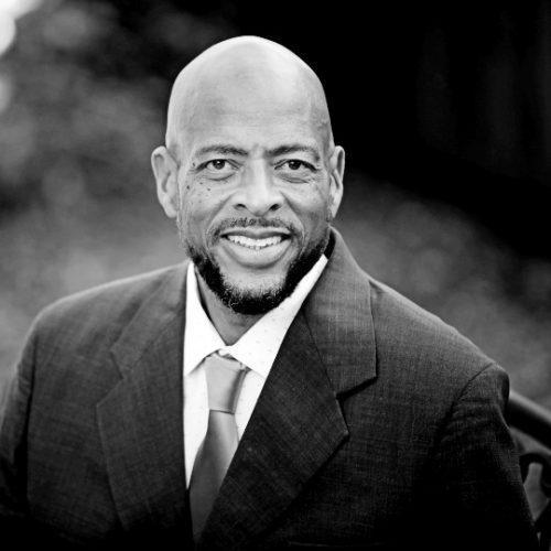 A person smiles while wearing a suit and tie, set against a blurred outdoor background in black and white.