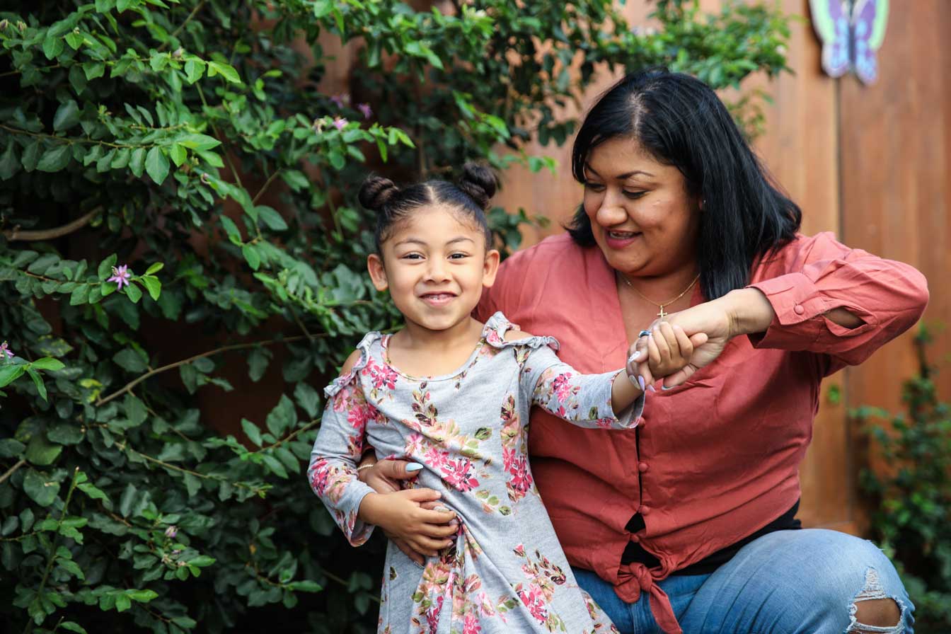 A child in a floral dress smiles while being playfully twirled by a woman in a red shirt, surrounded by lush green foliage in a garden setting.