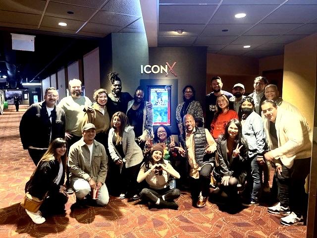 A group of people poses together in front of an illuminated poster on a wall labeled "ICON X," inside a carpeted indoor setting with overhead lighting.