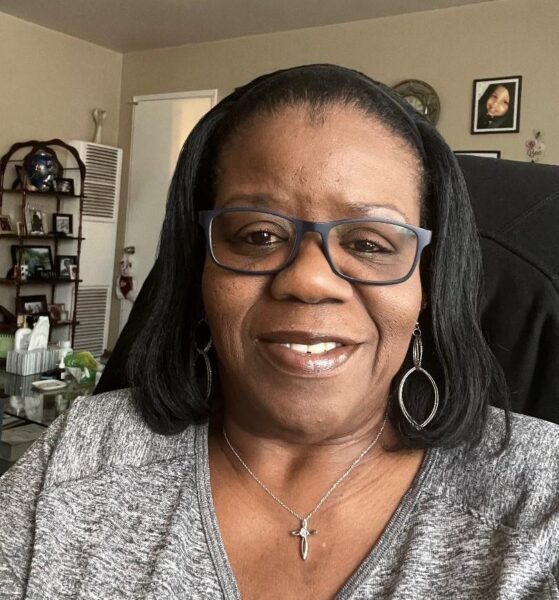 </p>
<h3>DeLoria White</h3>
<p> A woman with glasses and hoop earrings smiles at the camera, seated in a living room with framed pictures on the wall and a tall shelf in the background.