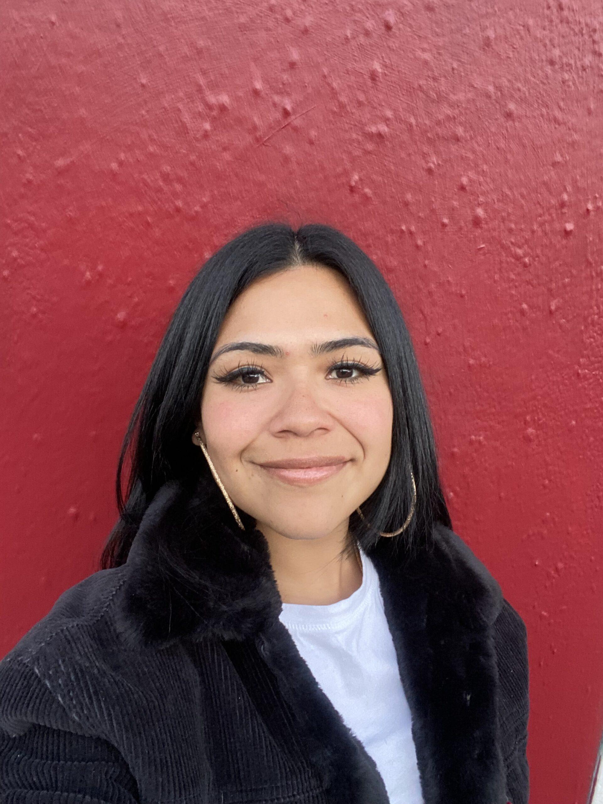 </p>
<h3>Dolores Del Carmen Ruiz</h3>
<p> A person smiles while standing against a textured red wall, wearing a black jacket and hoop earrings.
