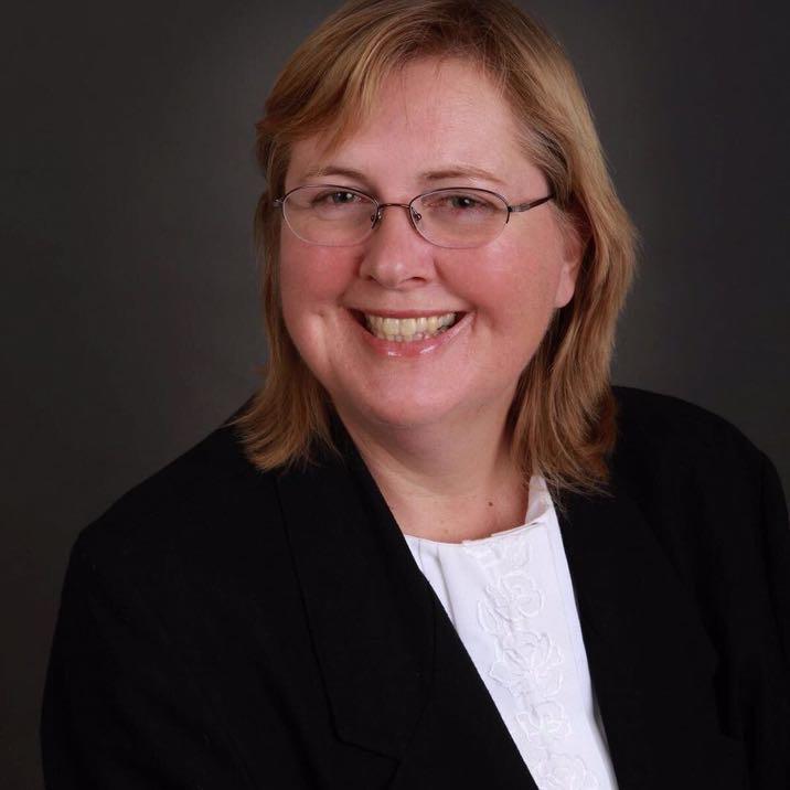 </p>
<h3>Sylvia Bambra</h3>
<p> A person with glasses smiles warmly, wearing a black blazer and white shirt, set against a dark, neutral backdrop.