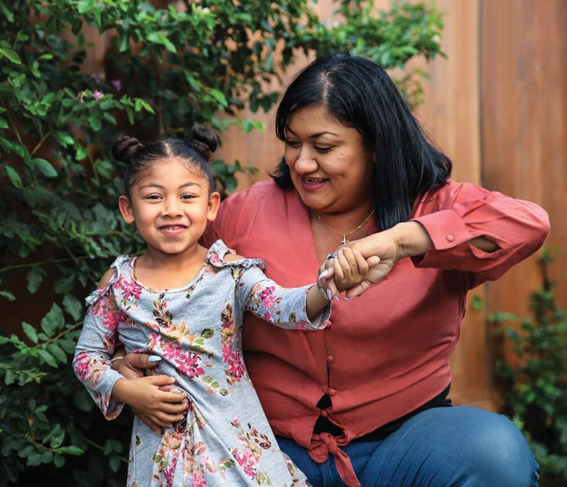 A woman in a red blouse gently holds the hand of a smiling young girl in a floral dress, sitting together near leafy greenery.