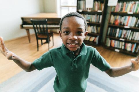 A smiling child in a green shirt stands with arms outstretched in a room with wooden floors, flanked by a piano on the left and bookshelves on the right.