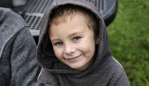 A young child, wearing a gray hoodie, smiles while sitting beside a metal picnic table; lush green grass is visible in the background.