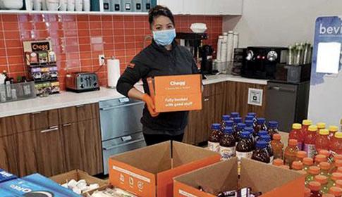 A person wearing a mask holds an orange Chegg box labeled “Fully furnished with good stuff” in a kitchen area filled with various juice bottles and packed food items.