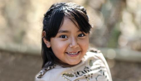A smiling young girl with dark hair wears a patterned shirt, standing outdoors in soft sunlight and blurred foliage background.