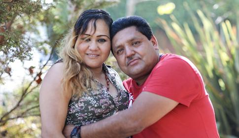 A couple embraces affectionately in a garden setting, surrounded by greenery and sunlight, conveying warmth and closeness.