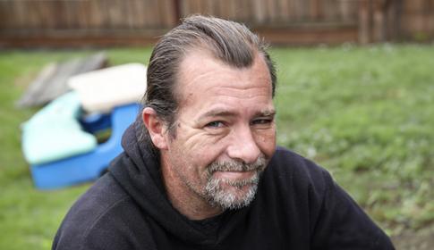 A person with short hair and a slight smile sits outdoors, wearing a dark hoodie. Wooden fencing and a patch of green grass occupy the background.