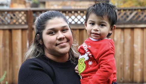 A person holds a smiling child wearing a red sweater with a Grinch design, standing outdoors in front of a wooden fence in a backyard setting.