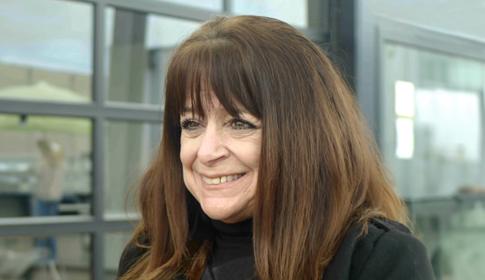 A woman with long brown hair smiles slightly, standing indoors in front of large windows, suggesting a modern or office environment.