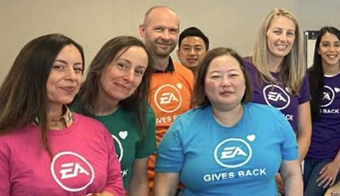 A group of people stands smiling, each wearing a colorful shirt with the text "EA GIVES BACK" and the EA logo. They are in a room with a neutral background.