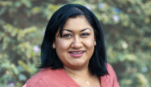 A person smiles directly at the camera, standing against a blurred background of green foliage.