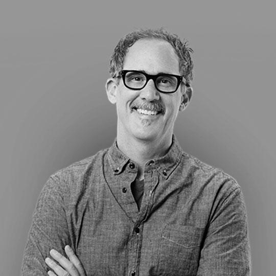 A person wearing glasses and a button-up shirt is smiling against a plain, neutral background.