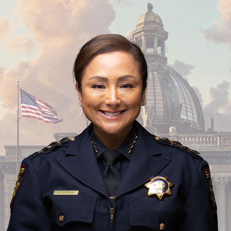 A person in a police uniform with a badge labeled "CORPUS" smiles in front of a backdrop featuring a domed building and an American flag on a cloudy day.