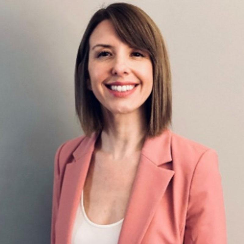 A person with shoulder-length hair is smiling while wearing a pink blazer over a white top against a plain, light-colored background.