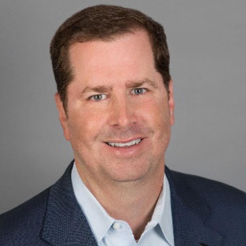 A man with short brown hair smiles slightly, wearing a blue blazer and light shirt, positioned against a plain gray background.