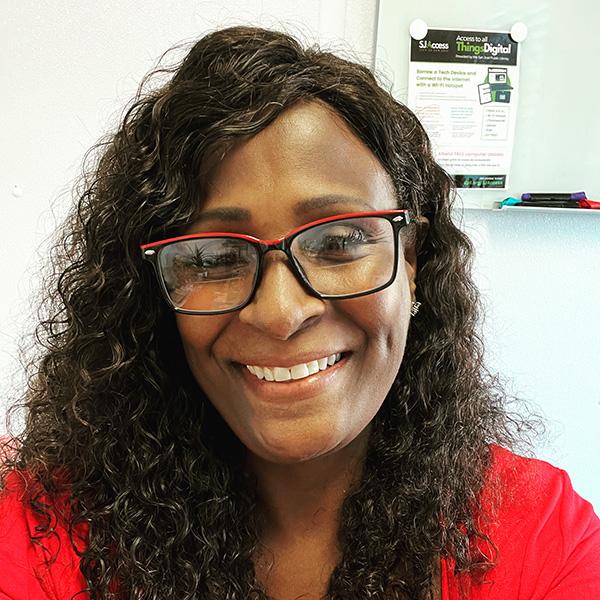 </p>
<h3>Toni Lynch</h3>
<p> A person with curly hair and glasses smiles at the camera, wearing a red shirt. A whiteboard with a flyer about "Access to All Things Digital" is visible in the background.