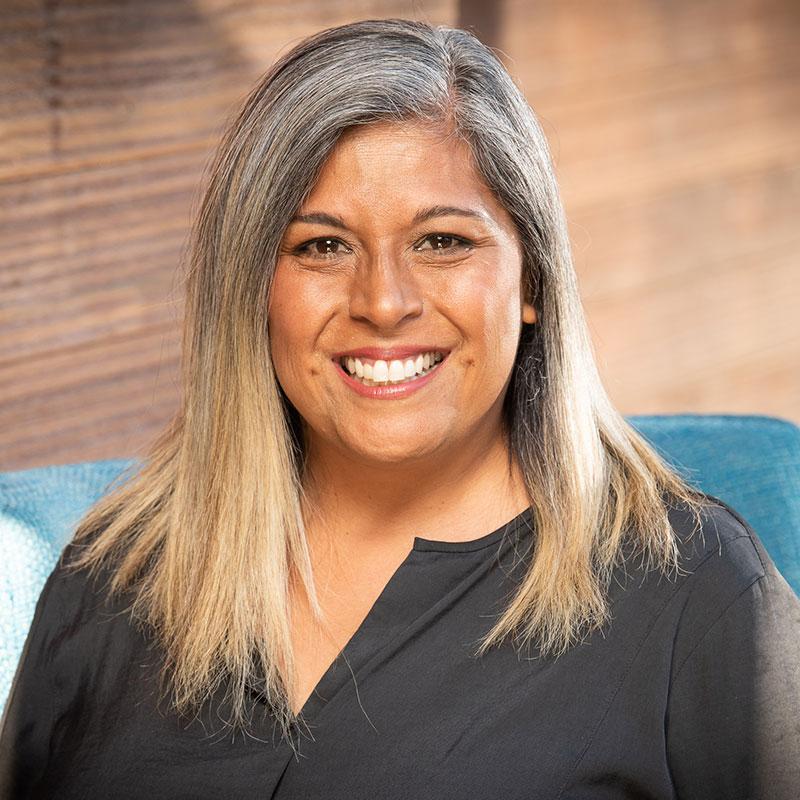 A person with gray hair smiles while sitting on a blue chair, framed by a wooden wall background.
