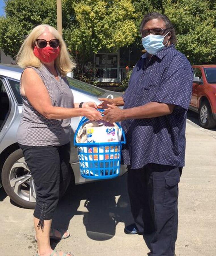 Two people wearing masks exchange a blue basket filled with groceries in a sunny outdoor parking lot. A gray car and trees are visible in the background.