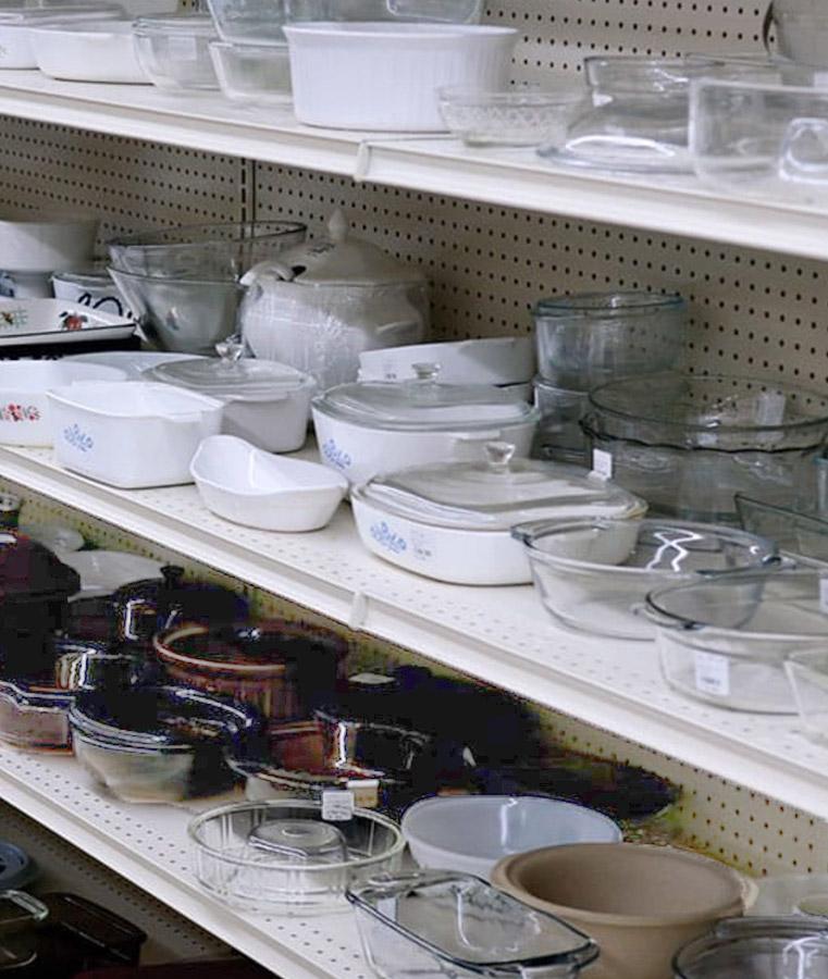 Glass and ceramic baking dishes lined up on store shelves, situated in a retail environment with pegboard backdrops. Various sizes and shapes are neatly organized, some with visible price tags.