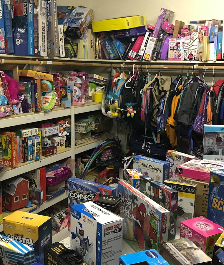 Shelves and tables filled with various toys and board games, such as "Connect 4," "Clue," and "Smart Robot," surround hanging backpacks in a cluttered storage room setting.