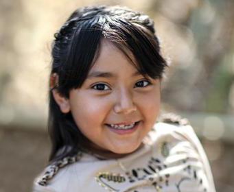 A smiling child stands outdoors, wearing a light-colored shirt with leopard print embellishments. The background is a blurred, sunlit natural setting.
