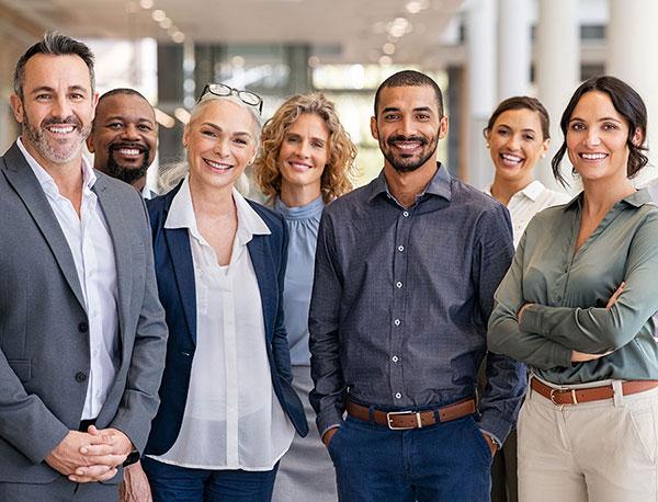 A group of seven smiling people stands together, posing confidently in business attire, within a spacious, modern office setting.