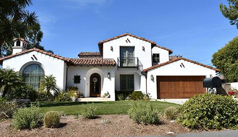 A two-story house with white stucco walls and red-tiled roof stands on a landscaped lawn, featuring arched windows and a wooden garage door, surrounded by greenery under a clear blue sky.