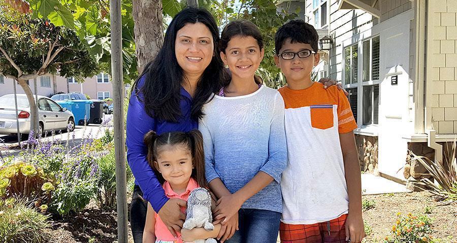 A woman and three children stand smiling in a garden. Trees and flowering plants surround them, with houses and parked cars visible in the background.