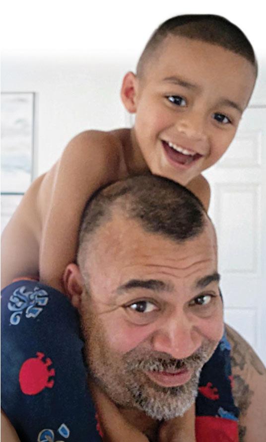 A child sits smiling on a man's shoulders in a bright room. The man, with a light beard, looks up playfully. Bright white walls and a partially visible door frame the scene.