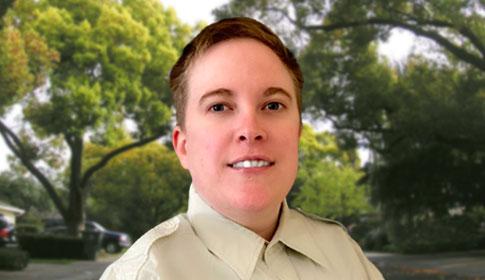 A person with short hair smiles slightly, wearing a light-colored shirt, standing outdoors with large, leafy trees in the background.