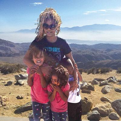 A person with three children stands embracing them on a rocky hillside, with distant mountains and a hazy sky in the background. The children wear colorful outfits.