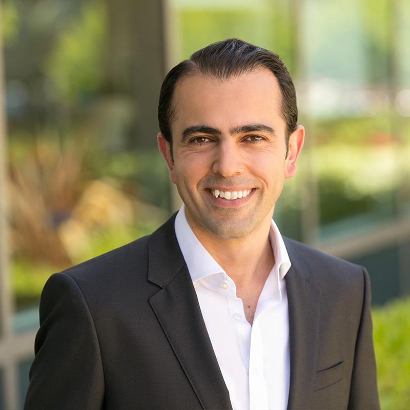 A man in a black suit and white shirt smiles warmly, standing outdoors with blurred greenery and glass windows in the background.